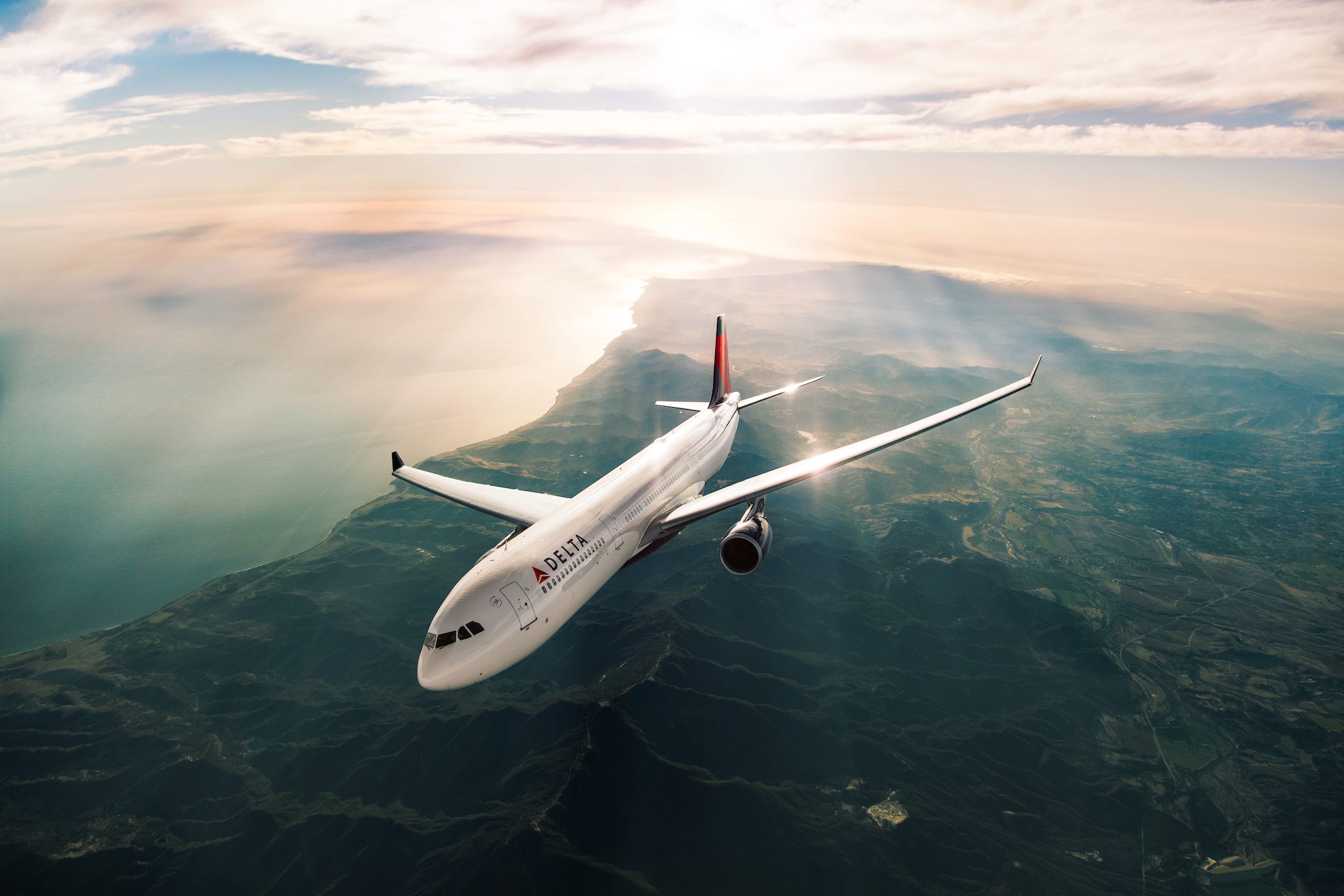 A Delta Air Lines Airbus A330 in flight climbing over a mountainous coastline with the sun shining in the background.