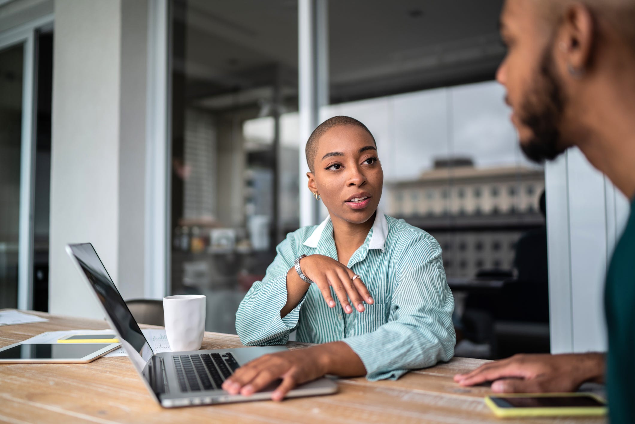 two people sitting at a desk talking with a laptop on the table between them