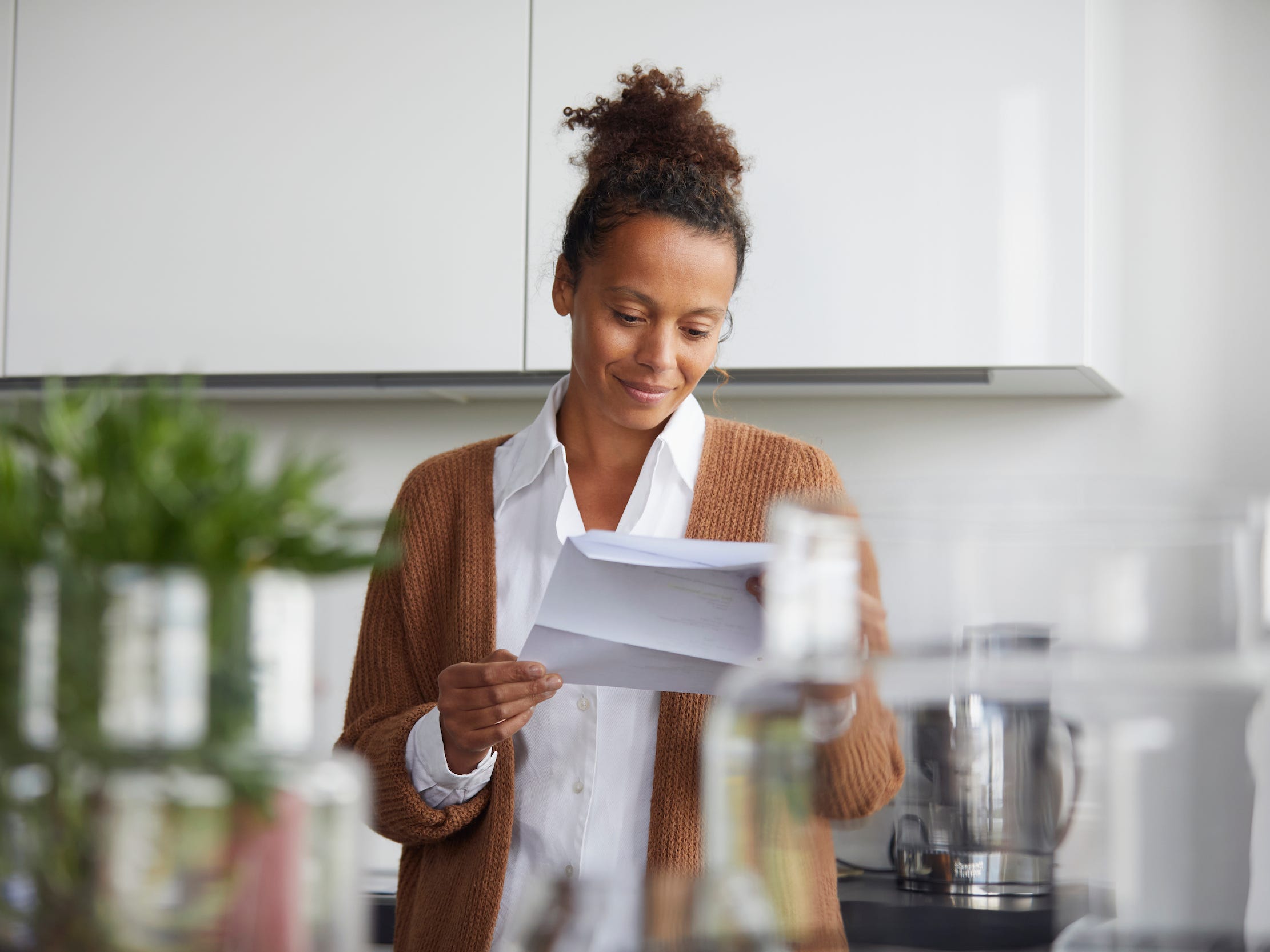 Smiling woman stands in kitchen with brown sweater reading a bill.