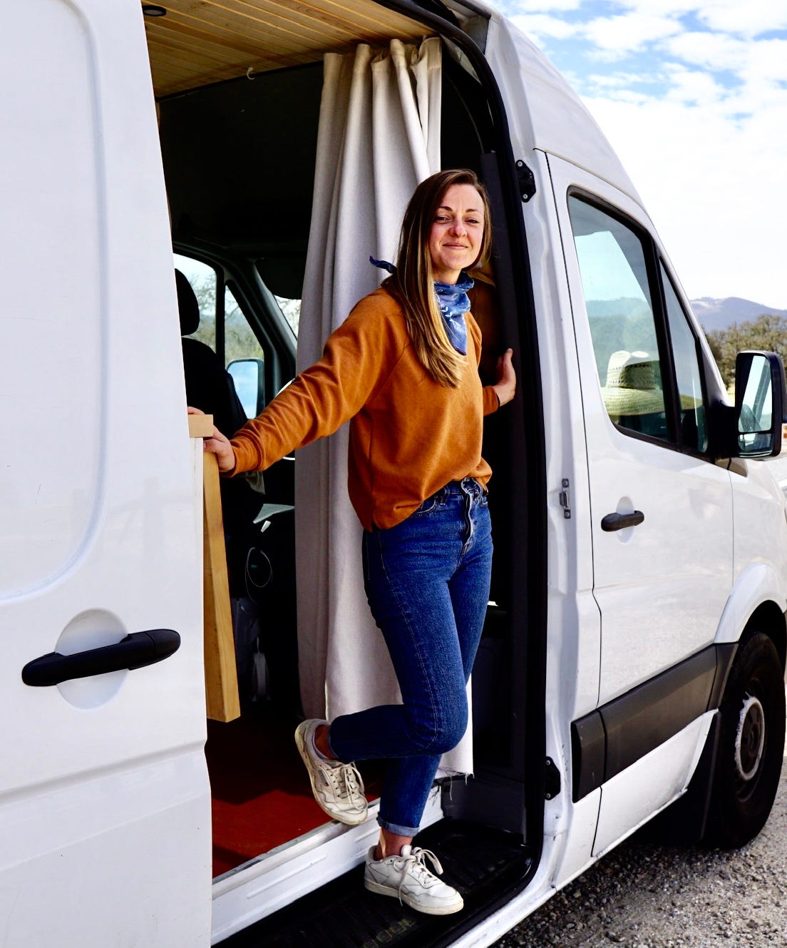 A smiling woman wearing an orange jumper stands in the doorway to her van.