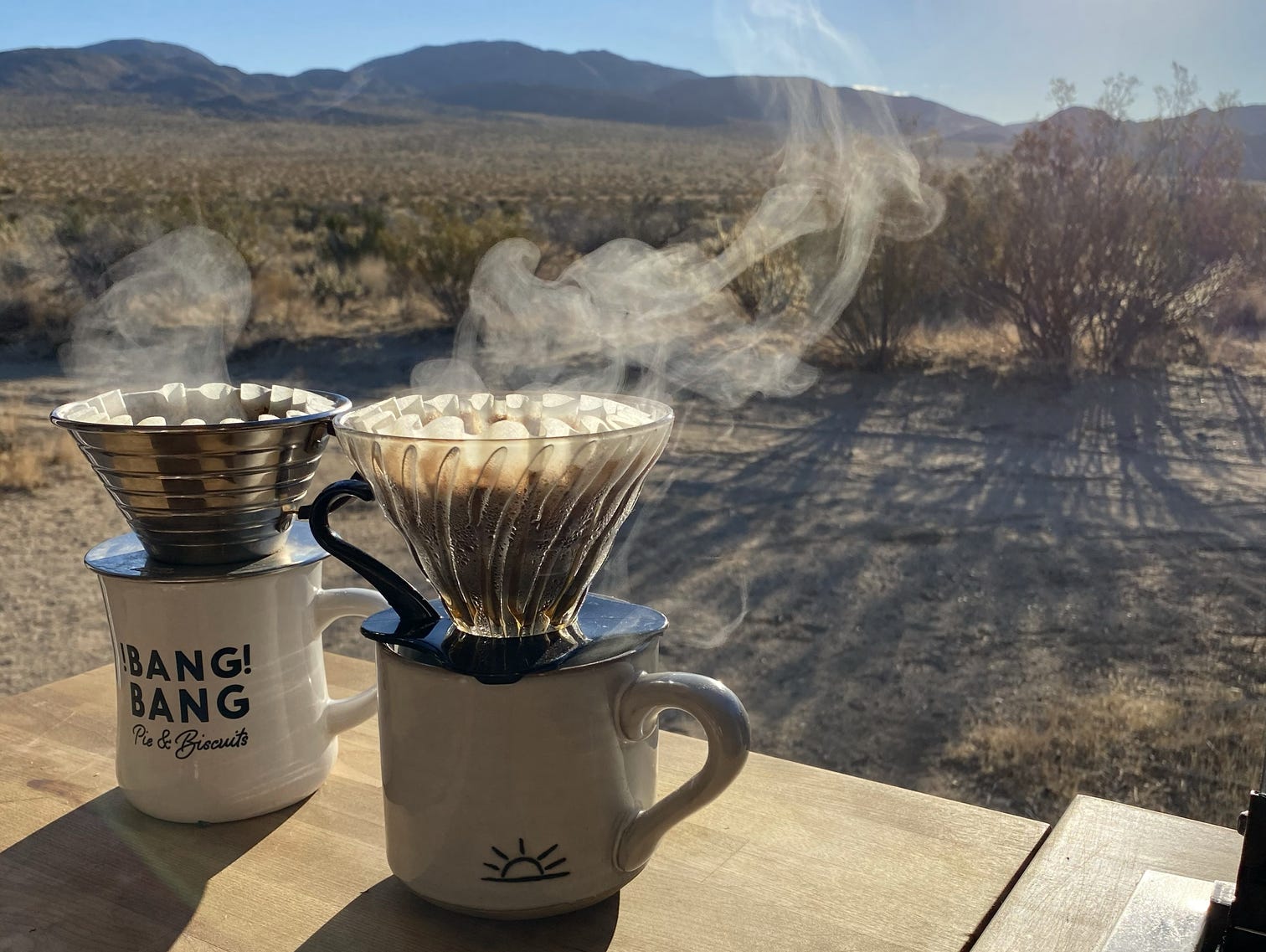 Two mugs containing hot drinks sit on a table with a mountainous scenery behind them.