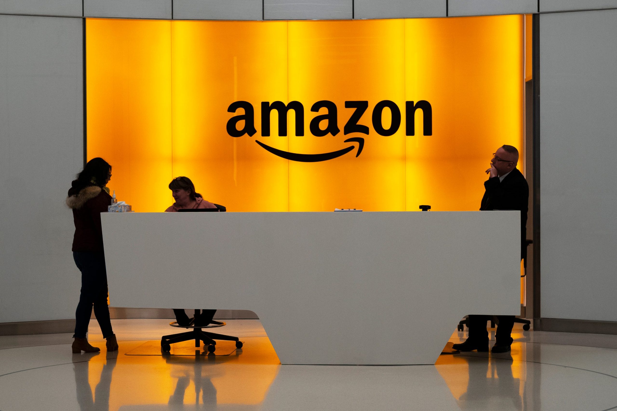 Amazon office front-desk staff stand in front of an orange sign with the Amazon smile logo